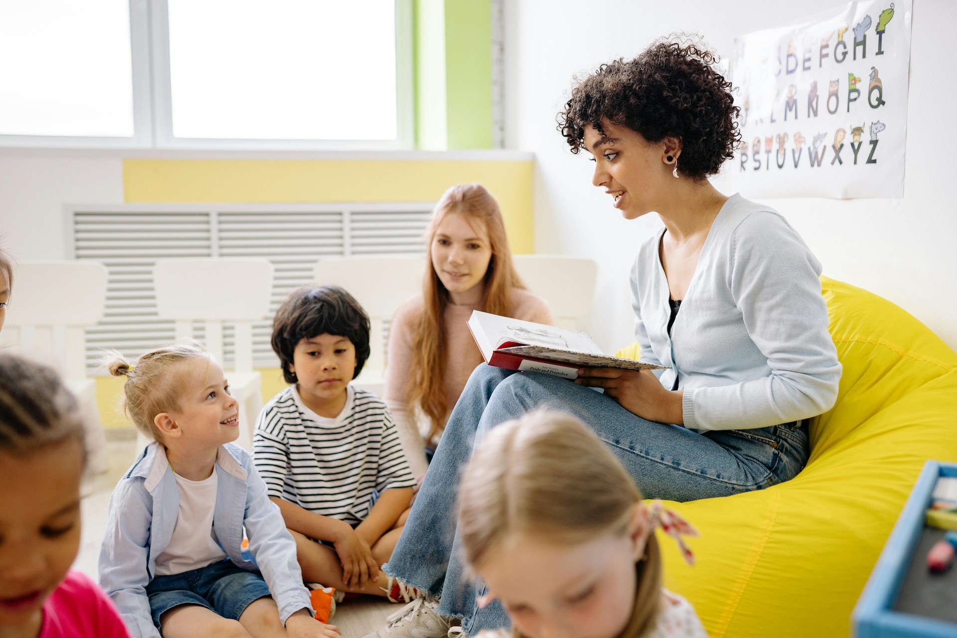 Pre-school teacher sitting on a chair holding a book with young children around them.