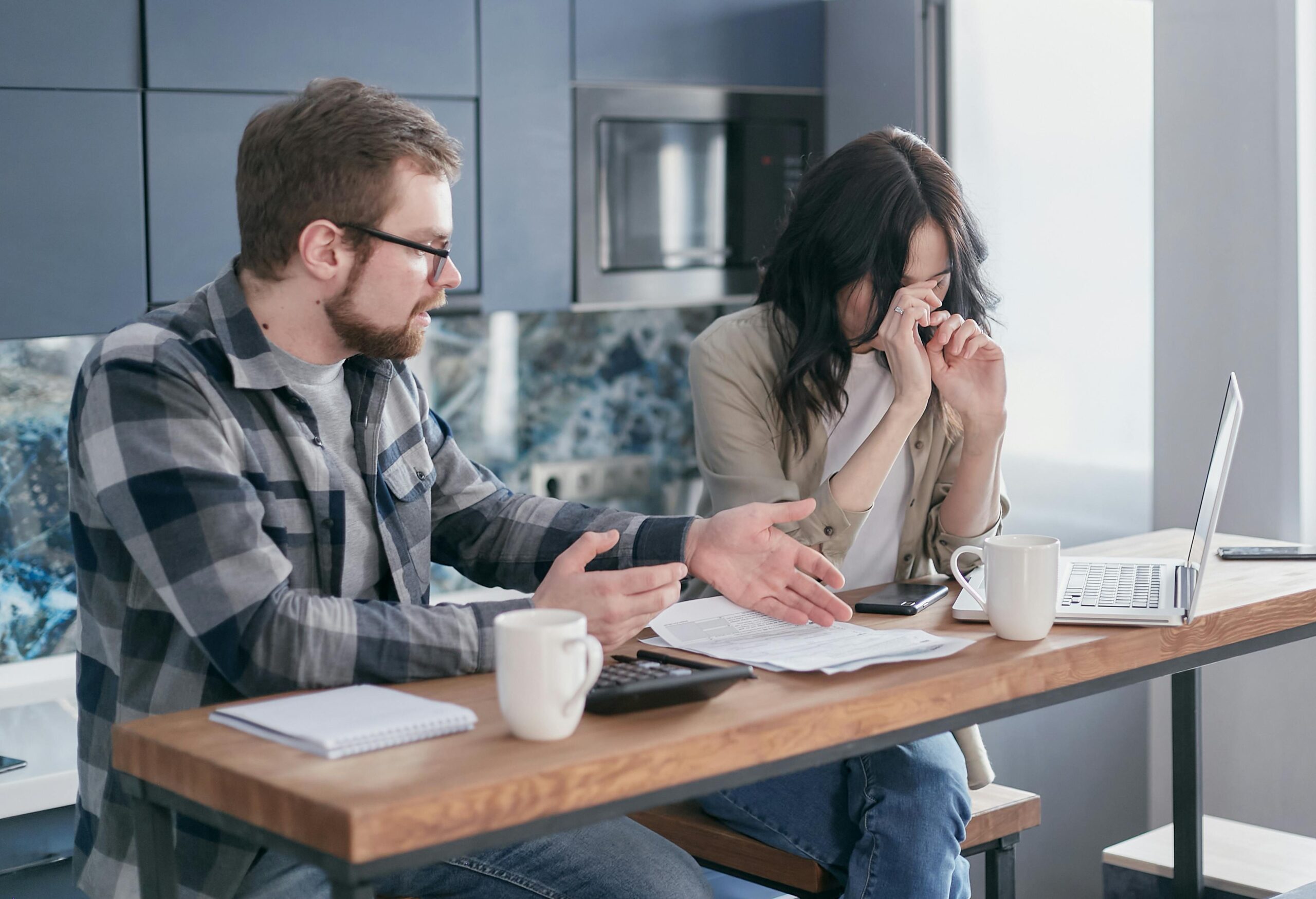 A husband and wife are sat at a table having a conversation. The husband is pointing to information on paper while the wife sits beside him looking distressed.