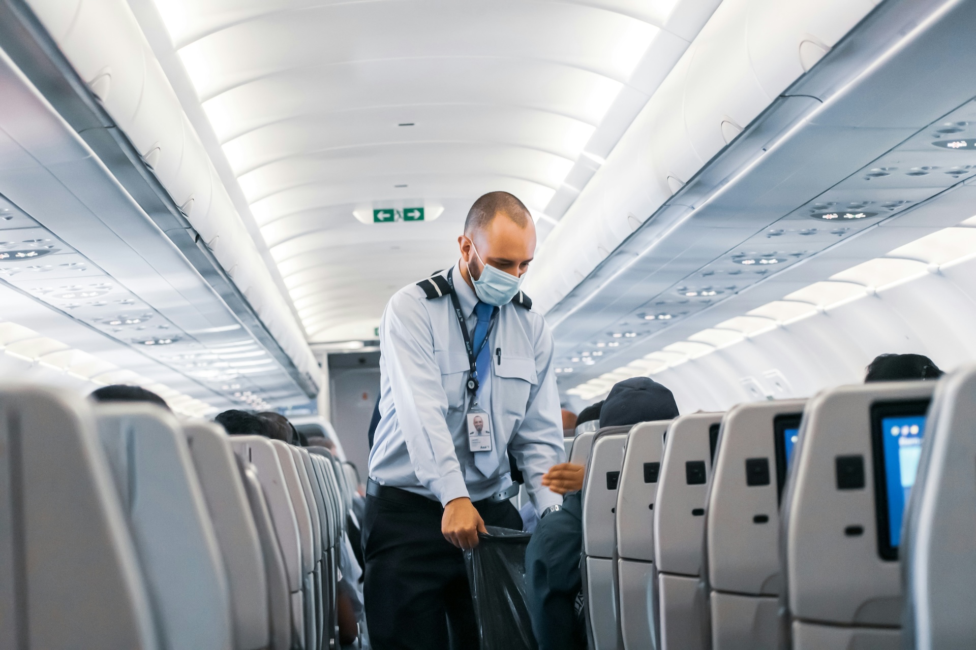 A male cabin crew member wearing a mask walking down the aisle of a plane holding a blue bag