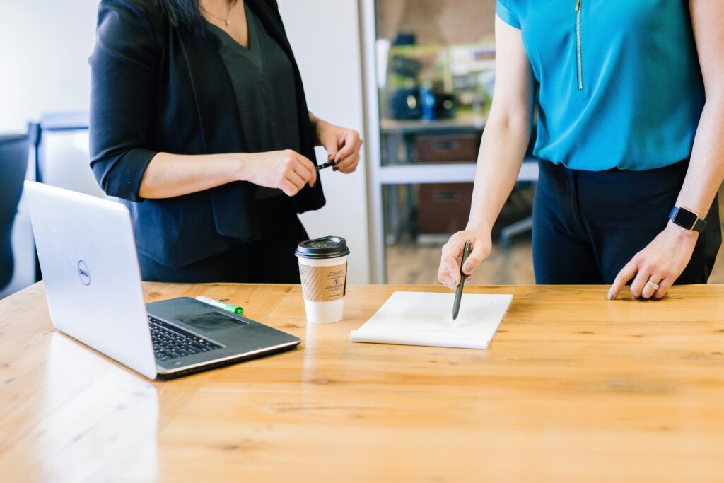 Two females standing behind a desk with a notepad, coffee cup and laptop on it. The female on the right who is wearing a blue shirt is holding a pen pointing to the notepad.